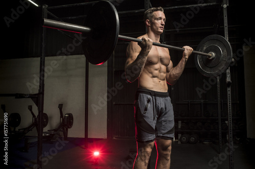 Muscular White Caucasian man doing a barbell bicep curl in a dark grungy gym wearing shorts and showing muscular body and six pack abs 