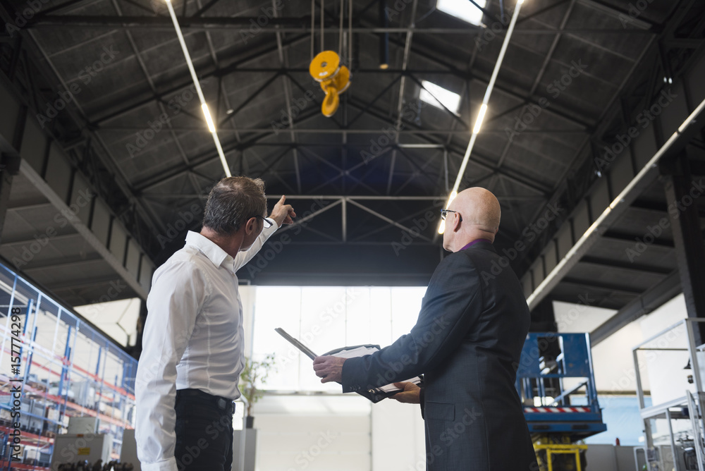 Two businessmen with clipboard on factory shop floor with bridge crane Two businessmen with clipboard on factory shop floor with bridge crane