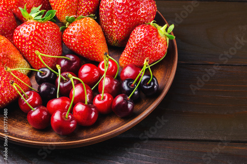 Harvest of fresh organic berry  strawberries and cherries in brown plate of clay on dark wooden planks background