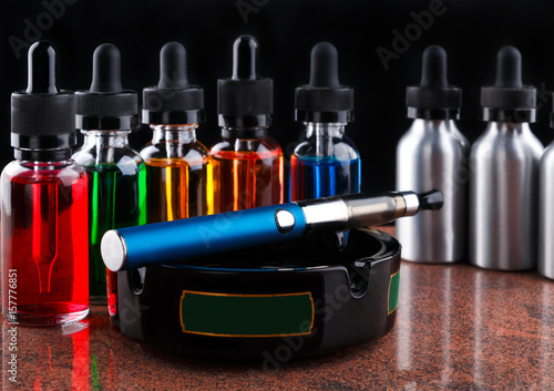 Electronic cigarette on the ashtray and bottles with vape liquid on black background photo