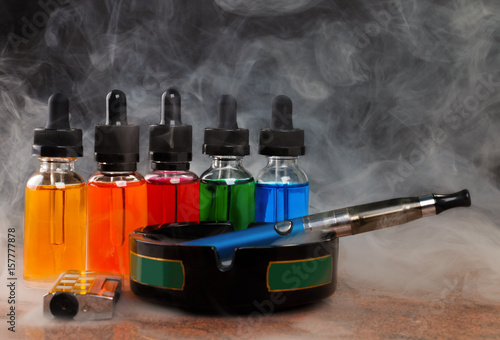 Electronic cigarette on ashtray, cigarette lighter and bottles with vape liquid within vapor on black background photo