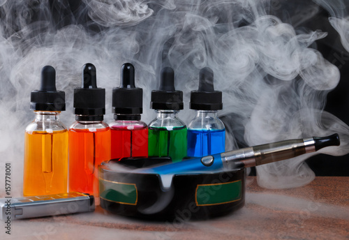 фотографія Electronic cigarette on ashtray, cigarette lighter and bottles with vape liquid