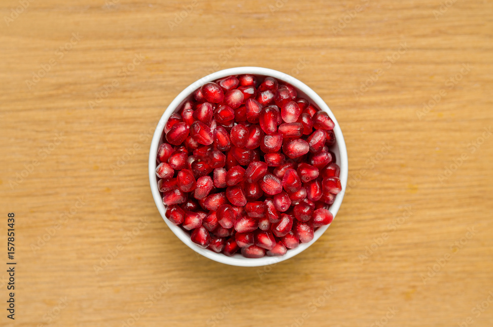 Grains Of Pomegranate In A White Bowl On A Wooden Board. Close-Up. Free Space For Text. Top View. In The Center. Grains Of Pomegranate In A White Bowl On A Wooden Board. Close-Up. Free Space For Text. Top View. In The Center.