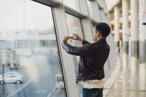 Handsome man standing beside glass wall in modern airport terminal, taking photo picture of airplane aircraft, travelling to other countires. photo