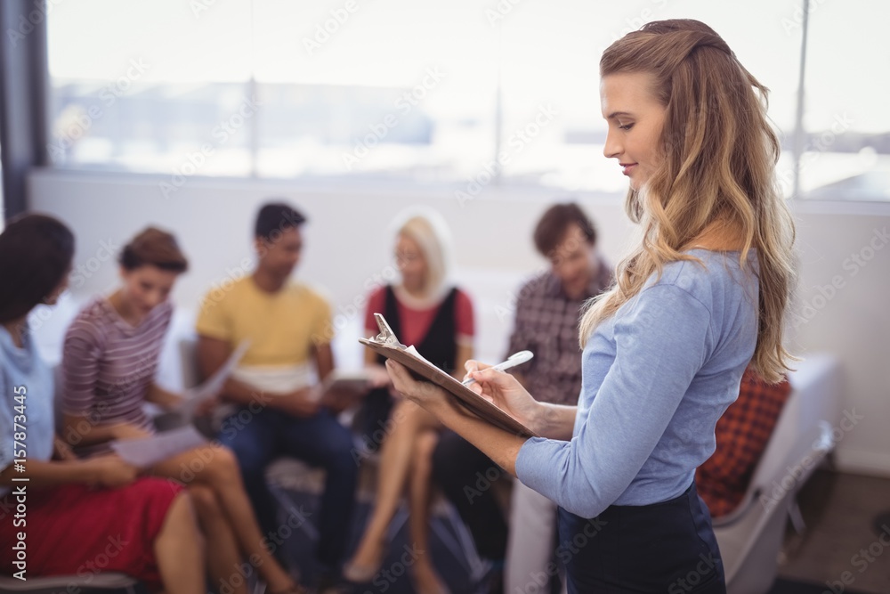 Businesswoman writing on clipboard with colleagues in background Businesswoman writing on clipboard with colleagues in background