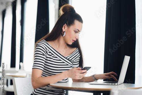 Charming Caucasian woman using mobile phone and laptop computer while sitting in modern coffee shop interior, young beautiful hipster girl holding cell telephone during work on portable net-book.