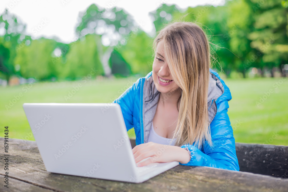 pretty smiling student chatting with friends on laptop in the summer park. pretty smiling student chatting with friends on laptop in the summer park.