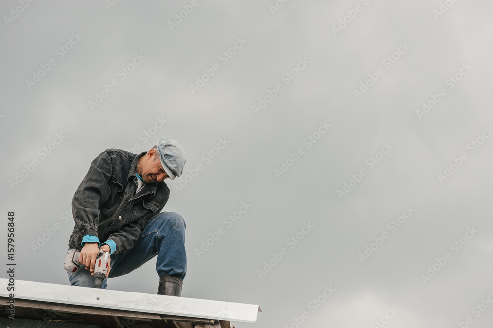 A man in work clothes tighten screws with a screwdriver on the roof against a cloudy sky. A man in work clothes tighten screws with a screwdriver on the roof against a cloudy sky.