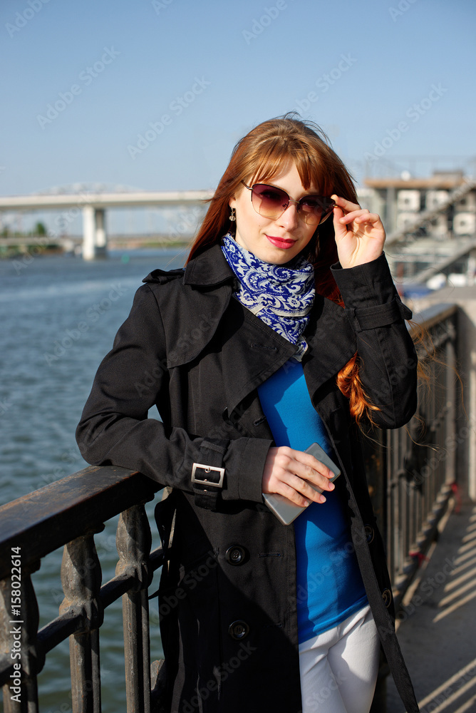 Beautiful young woman leaned against the railing on the embankment, holding a mobile phone and corrects sunglasses.