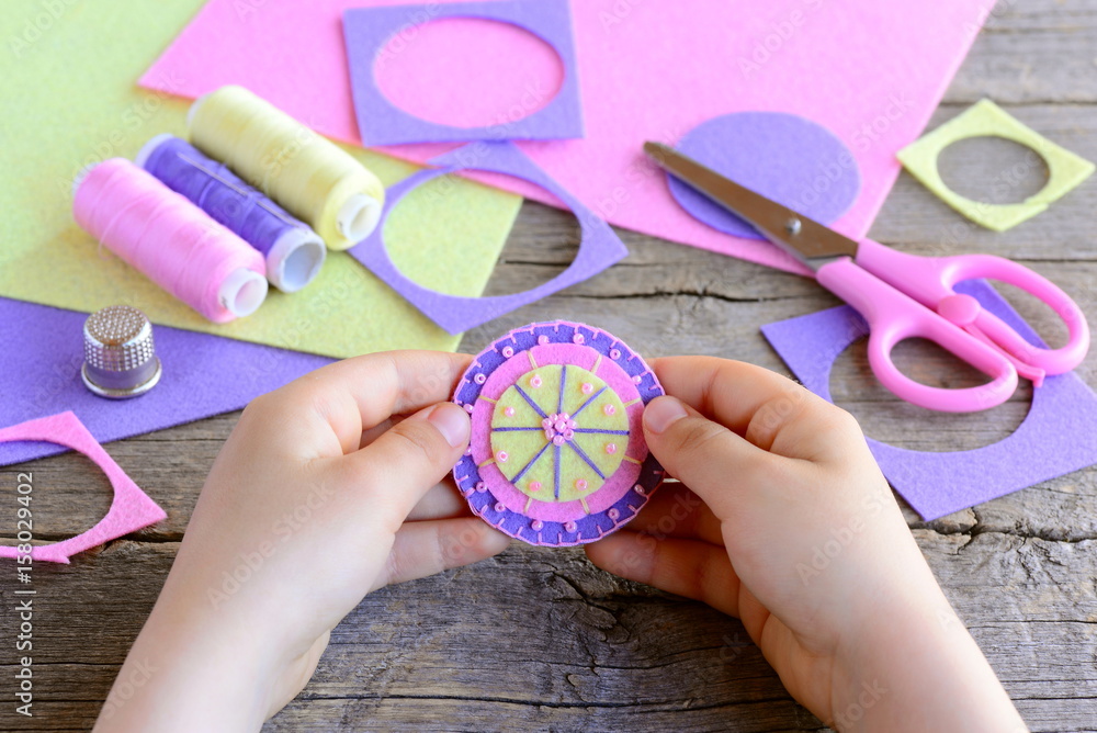 Fototapeta premium Small child made a simple and colorful flower from felt circles and beads. Child holds a bright felt flower in hands. Craft supplies on wooden table. Easy and fun teaching small kids to sew by hand