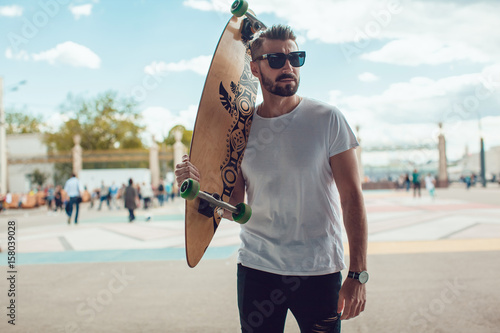 Stylish man in sunglasses and with a beard stands on the street with a long board. T-shirt mock up. photo