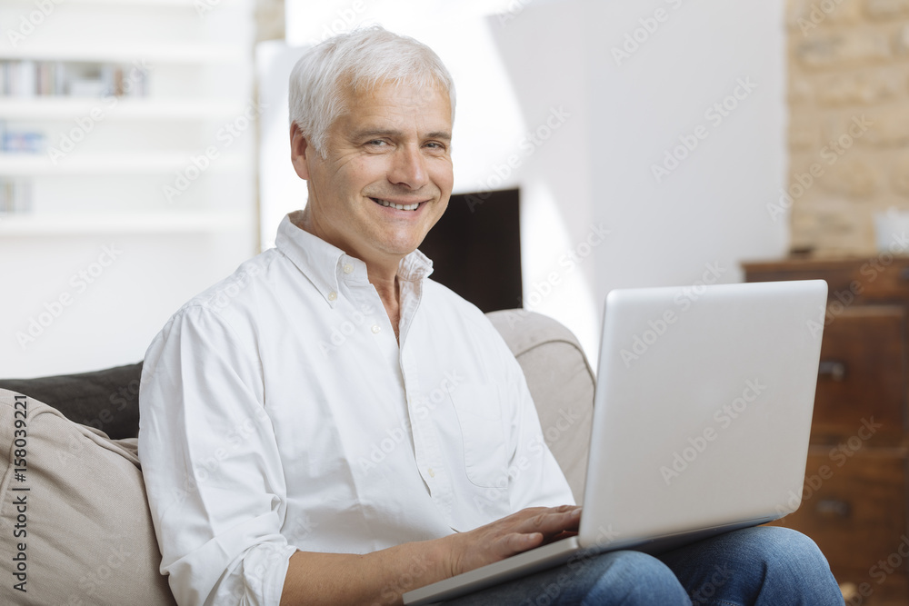 Smiling mature man sitting on a sofa typing on a laptop in a living room Smiling mature man sitting on a sofa typing on a laptop in a living room