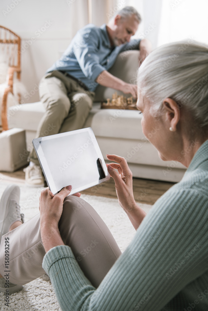 smiling senior woman using digital tablet while her husband playing chess on sofa smiling senior woman using digital tablet while her husband playing chess on sofa