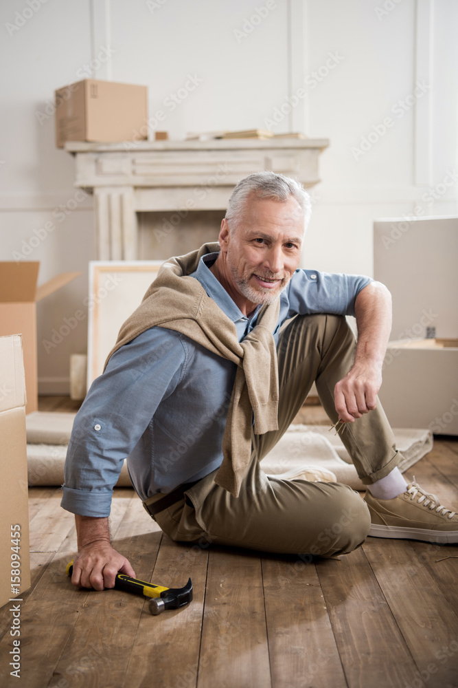 smiling senior man sitting on floor with hammer in hand at new home smiling senior man sitting on floor with hammer in hand at new home