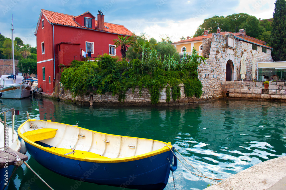 Cozy picturesque town of Zadar, Croatia. Red house among green ivy foliage near emerald water where one blue and yellow boat and several yachts are floating. Dramatic cloudy morning sky