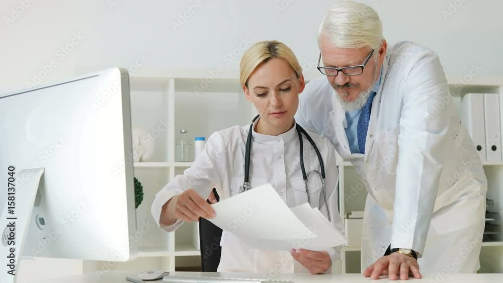 Caucasian young female doctor talking about something looked on sheets op paper with caucasian midle aged male doctor. Indoor. Caucasian young female doctor talking about something looked on sheets op paper with caucasian midle aged male doctor. Indoor.