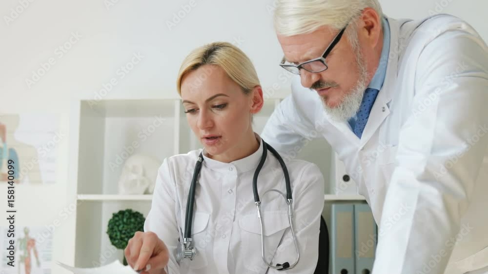 Portrain of caucasian female doctor sitting in her office, talking about something looked on sheets op paper with caucasian male doctor. Indoor. Portrain of caucasian female doctor sitting in her office, talking about something looked on sheets op paper with caucasian male doctor. Indoor.