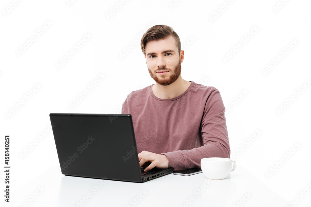 Handsome young man at the table looking in the camera working at the laptop computer isolated Handsome young man at the table looking in the camera working at the laptop computer isolated