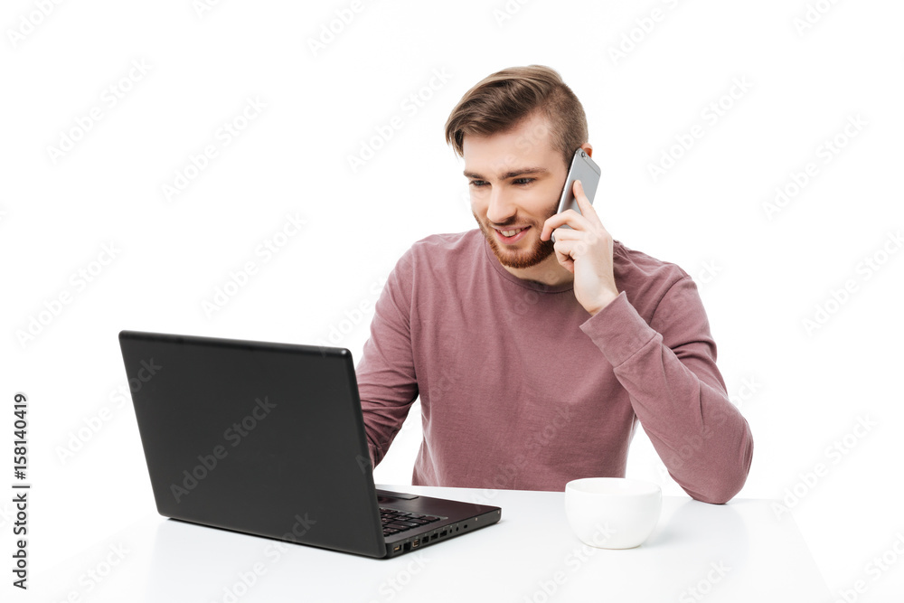 Handsome young man working at the laptop computer talking on the phone and looking at the display isolated Handsome young man working at the laptop computer talking on the phone and looking at the display isolated