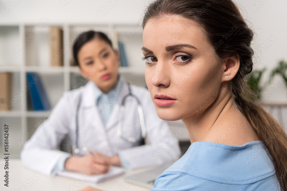 portrait of upset patient looking at camera with doctor sitting at workplace behind portrait of upset patient looking at camera with doctor sitting at workplace behind