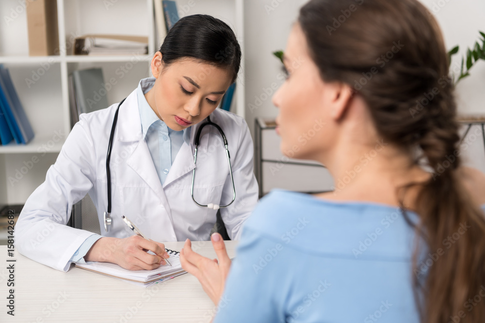 doctor writing in notebook while patient telling about illness at hospital