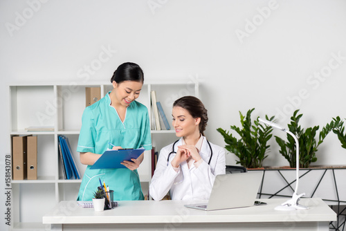 smiling doctor using laptop and talking with young asian internist writing in clipboard in clinic