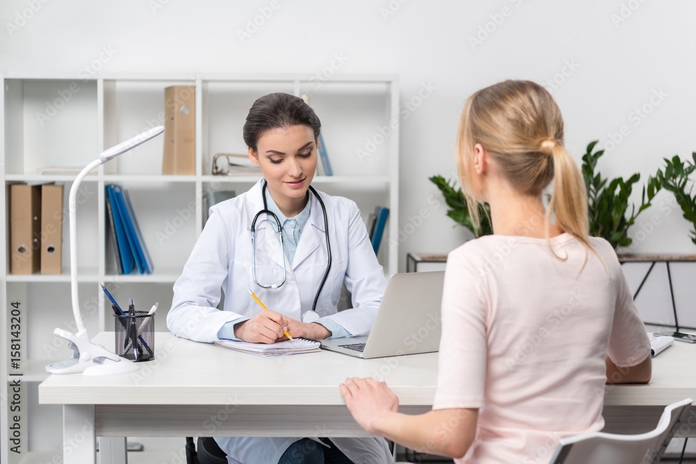 Young doctor with stethoscope taking notes while talking with patient in clinic Young doctor with stethoscope taking notes while talking with patient in clinic