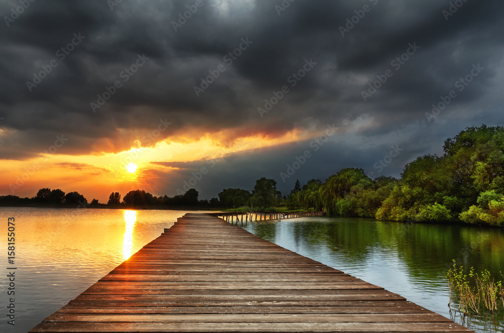 Fototapeta Wooden path bridge over lake at stormy dramatic sunset