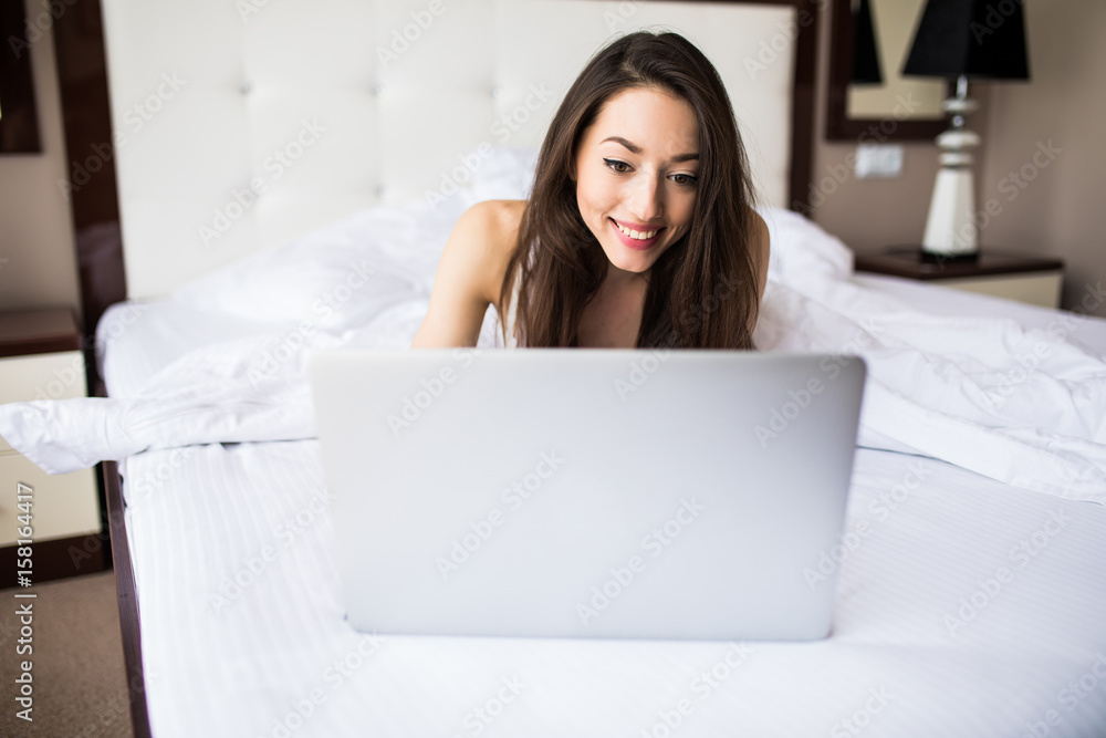 Smiling woman catching up on her social media as she relaxes in bed with a laptop computer on a lazy day Smiling woman catching up on her social media as she relaxes in bed with a laptop computer on a lazy day