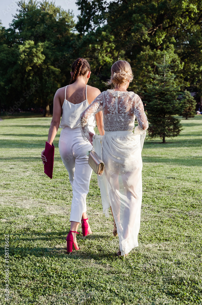 two elegant women with their backs walking through the park on the grass two elegant women with their backs walking through the park on the grass