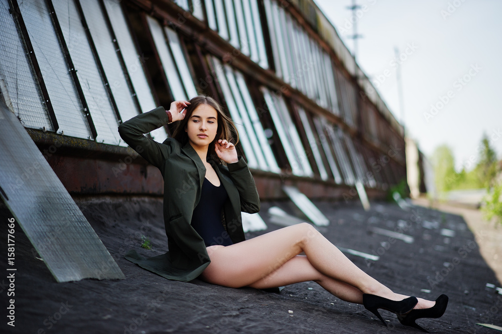 Sexy model girl wit long legs at black lingerie outfit body swimsuit combidress and jacket posed at the roof of abadoned industrial place with windows.