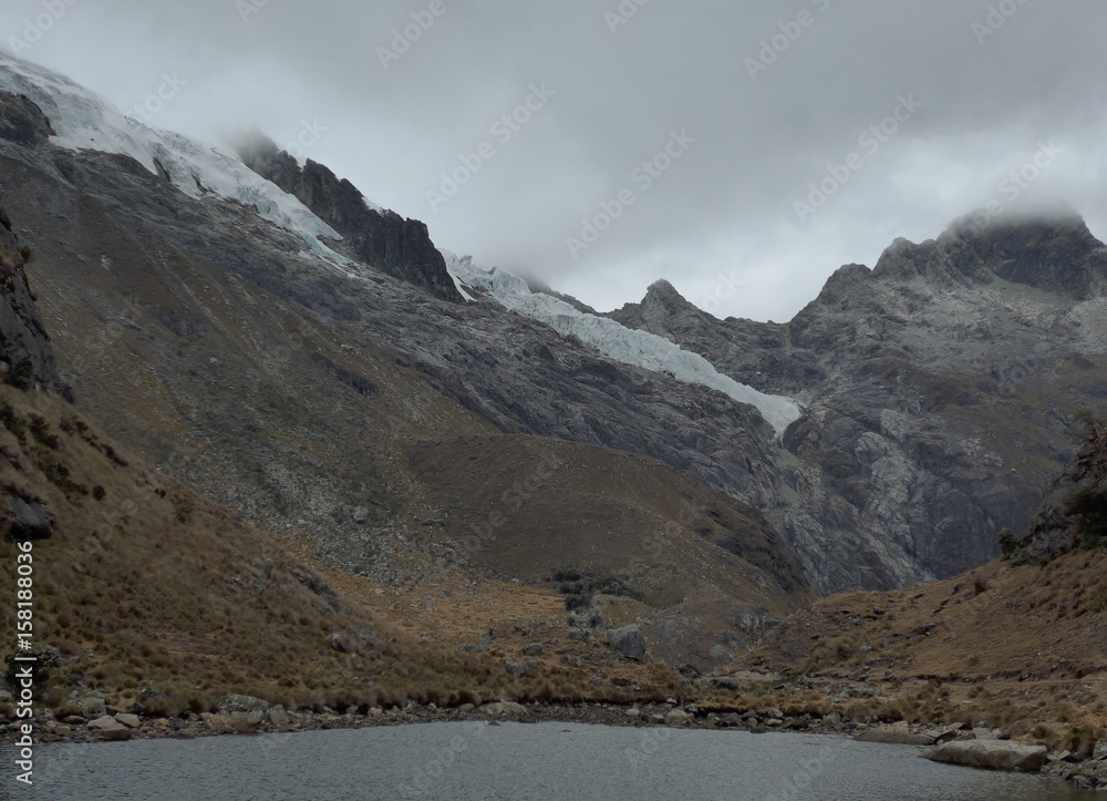Dark overcast day, glaciers hanging on to the top of a dark grey rocky mountains side above the cold waters of an alpine lake 