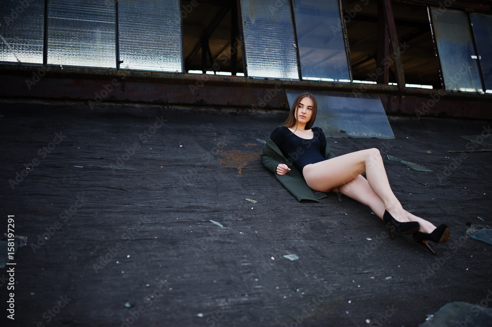 Sexy model girl wit long legs at black lingerie outfit body swimsuit combidress and jacket posed at the roof of abadoned industrial place with windows.