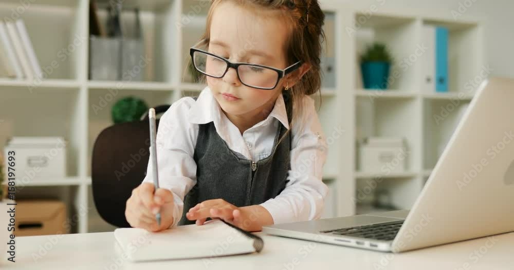 Serious young girl with glasses and brown hair writing something in notebook while typing on the laptop siting on the desk in office. Indoor. Serious young girl with glasses and brown hair writing something in notebook while typing on the laptop siting on the desk in office. Indoor.