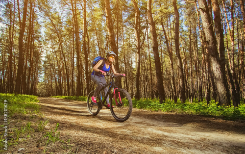 One young woman - cyclist in a helmet riding a mountain bike outside the city, on the road in a pine forest on a summer day.