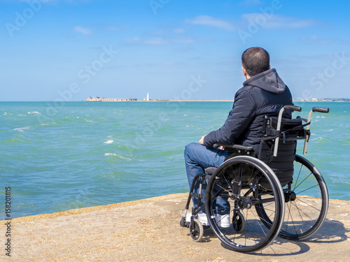 Disabled young man sitting in a wheelchair and looks at the sea.