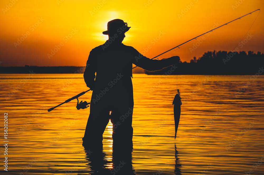 A fisherman with a fishing rod in his hand and a fish caught stands in the water against a beautiful sunset. A fisherman with a fishing rod in his hand and a fish caught stands in the water against a beautiful sunset.