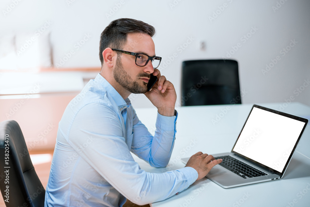 Attractive european guy talking on phone while using laptop at workplace Attractive european guy talking on phone while using laptop at workplace