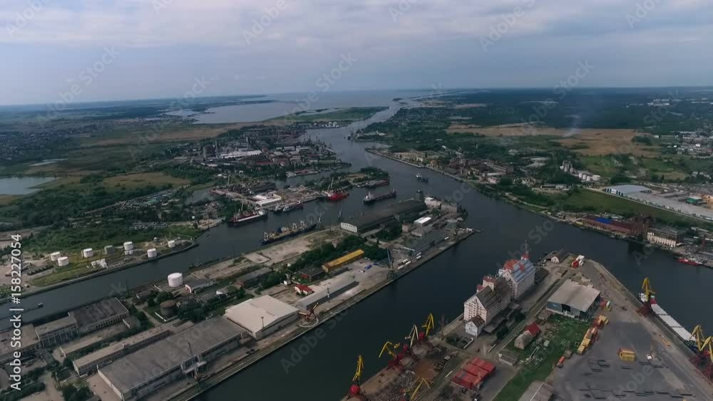 Sea Port. Concrete Banks. Ships in Bay. Cranes Loading Boats. Green Trees by City. Storage Buildings Warehouse on Shore. Forest on Background. Summer Sea Port. Concrete Banks. Ships in Bay. Cranes Loading Boats. Green Trees by City. Storage Buildings Warehouse on Shore. Forest on Background. Summer