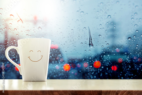 Happy Coffee Mug with smilely face on desk inside glass window, Blurred traffic jam light in city as outside view, Relaxing in cafe on rainy day photo