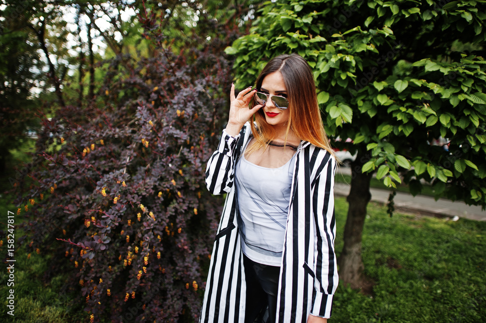 Fashionable woman look with black and white striped suit jacket, leather pants and sunglasses posing against bushes at street. Concept of fashion girl. Fashionable woman look with black and white striped suit jacket, leather pants and sunglasses posing against bushes at street. Concept of fashion girl.