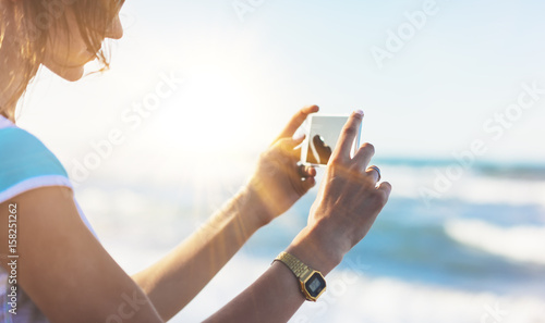 Hipster girl photograph on smart phone gadget in sand coastline, mock up of blank screen. Traveler using in female hand mobile on background seascape horizon. Tourist look on blue sun ocean, lifestyle
