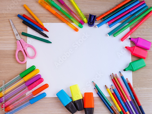 A white sheet of paper lay on a wooden table, near , pencils, markers, pens . The view from the top.