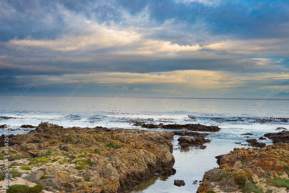 Rocky coast line on the ocean at De Kelders, South Africa, famous for whale watching. Winter season, cloudy and dramatic sky. Rocky coast line on the ocean at De Kelders, South Africa, famous for whale watching. Winter season, cloudy and dramatic sky.