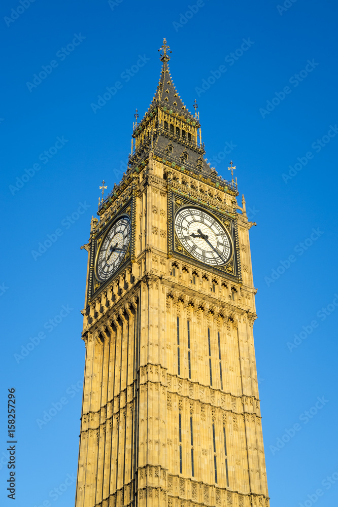 The Gothic architecture of the Big Ben clocktower, officially known as Queen Elizabeth Tower, shining golden in summer sunset light against bright blue sky