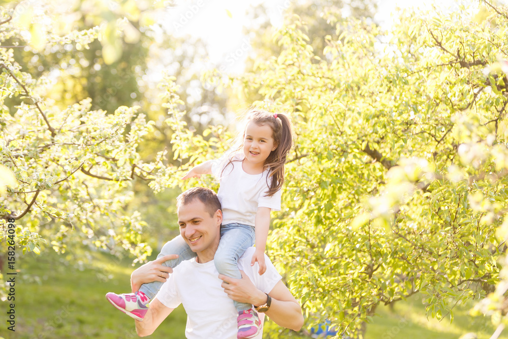 Attractive young man is playing with his daughter in the nature. The father is standing and carrying girl on his back. He is stretching arms sideways. The family is smiling Attractive young man is playing with his daughter in the nature. The father is standing and carrying girl on his back. He is stretching arms sideways. The family is smiling