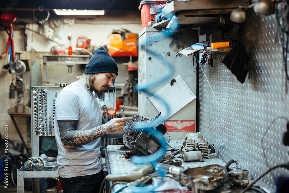 Side view portrait of modern tattooed man fixing broken parts at table in workshop Side view portrait of modern tattooed man fixing broken parts at table in workshop