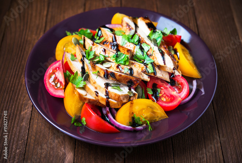 Sliced roasted chicken breast and fresh salad with tomato and purple onion topped with balsamic mousse, pumpkin seed and parsley close up on dark wood background. Healthy food.