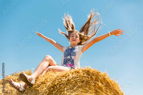 Young happy girl with long blond hair sitting on haystacks in a field of ripe wheat.