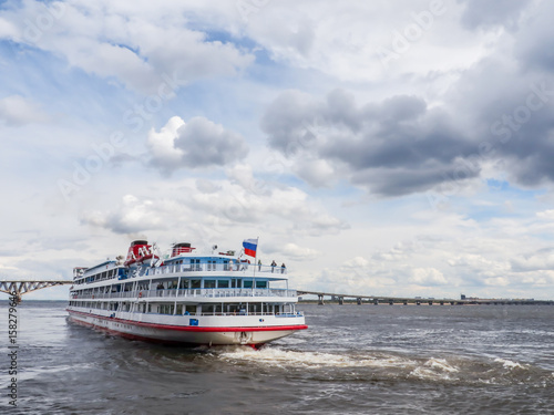 Four-deck passenger ship departs from the pier of the city of Saratov. On the horizon road bridge. The Volga River, Russia. White clouds on a blue sky.